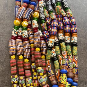 Puede incluir: Una colección de coloridas cuentas de vidrio hechas a mano. Las cuentas presentan una variedad de patrones, incluyendo rayas, espirales y diseños florales, en tonos de rojo, azul, amarillo, verde y blanco. Las cuentas están ensartadas con un cordón de fibra natural.