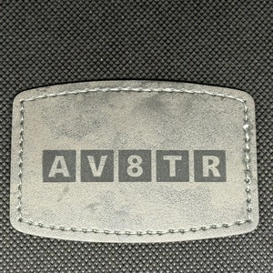 May include: A gray leather patch with the text "AV8TR" in black lettering.