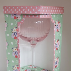 Könnte beinhalten: Ein rosa Glas mit einem Blumenmuster auf der Schachtel. Die Schachtel ist aus Pappe und hat ein weißes Punkte-Muster auf der Oberseite.