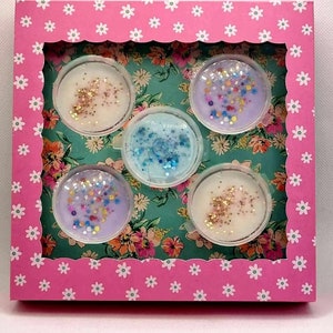 Può includere: Una scatola rosa con un bordo a festoni e un motivo a margherita bianca. All'interno della scatola ci sono sei piccoli contenitori rotondi con cere profumate di diversi colori. Le cere profumate hanno glitter incorporati.
