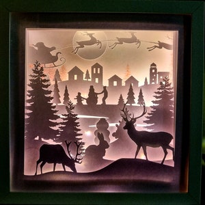 Puede incluir: Una caja de sombras con temática navideña con un diseño en capas. La escena representa siluetas del trineo de Papá Noel, renos, casas, árboles y ciervos. La obra de arte está iluminada desde dentro, creando un brillo cálido y festivo.