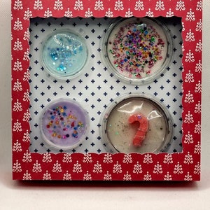 Könnte beinhalten: Eine rote Box mit einem weißen Weihnachtsbaum-Muster enthält vier Wachs-Schmelzen. Die Schmelzen befinden sich in durchsichtigen Behältern mit verschiedenen Farben und Designs, darunter blauer Glitzer, rosa Glitzer und ein roter Zuckerstock.