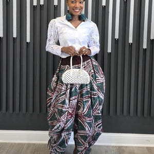 May include: A woman wearing a white eyelet shirt, patterned pants in shades of green, burgundy, and white, and white pointed-toe shoes. She is holding a white handbag with a dotted pattern. The background features a gray and white striped wall.