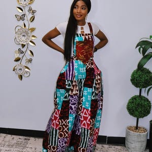 Op de afbeelding: Een vrouw draagt een patchwork jumpsuit met daaronder een wit t-shirt. De jumpsuit heeft een verscheidenheid aan kleurrijke patronen, waaronder geometrische vormen en symbolen. De merknaam "SamandaRose" staat in gouden letters op de muur.