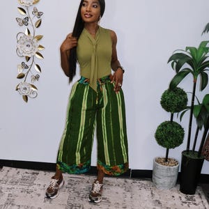 Peut inclure: Un haut sans manches vert olive avec un détail de nœud, associé à un pantalon large à rayures vertes. Le pantalon a un motif floral coloré à l'ourlet. La tenue est accessoirisée avec des baskets. Le mur derrière la tenue porte le texte "Samanda Rose".