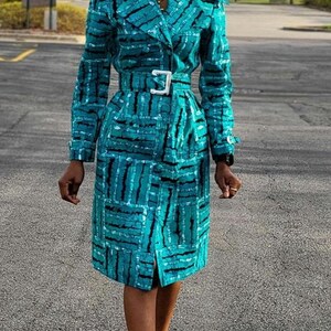 May include: A teal blue patterned trench coat with a white belt. The coat has a long sleeve and a collar. The model is wearing black heels.