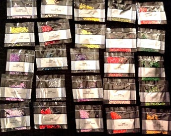 Può includere: Una collezione di piccoli pacchetti sigillati pieni di perline colorate. Ogni pacchetto ha un'etichetta bianca con il testo "Forever Young" e un codice prodotto. Le perline sono di vari colori, tra cui giallo, rosso, verde e viola.