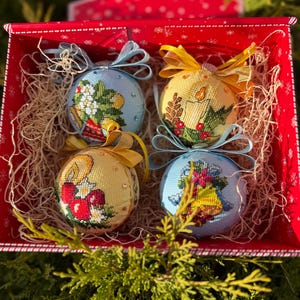 May include: Four embroidered Christmas ornaments in a red box with decorative straw. The ornaments are round, with blue and yellow backgrounds, and feature embroidered designs of candles, flowers, and bells. Each ornament has a ribbon for hanging.
