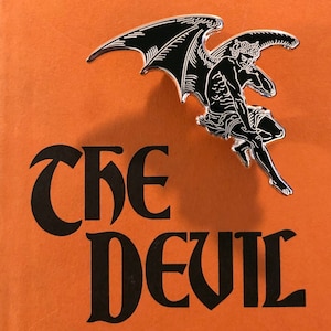 May include: A silver pin featuring a black and white image of a devil with wings. The text "THE DEVIL" is printed on an orange background.