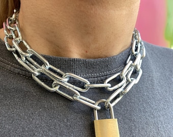 May include: A silver-colored chain necklace with a gold-colored padlock pendant. The necklace is made of large, interlocking chain links. The padlock is rectangular and hangs from the center of the chain.
