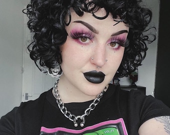 May include: A person with black curly hair, wearing a black t-shirt with a graphic of a vintage television displaying a green image. They have dark lipstick, pink eyeshadow, and a silver chain necklace.