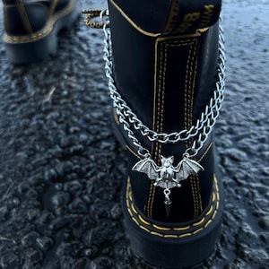 Może przedstawiać: Czarne skórzane buty z żółtymi przeszyciami i srebrnym łańcuchem z nietoperzem. Zawieszka z nietoperzem ma rozłożone skrzydła i zwisający kolec. Buty znajdują się na ciemnej, teksturowanej powierzchni.