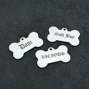 May include: Three silver dog tags in a bone shape, each with a small hole for attachment. The tags are engraved with the words "Dani", "Soft Boy", and "Vicious" in a decorative font.