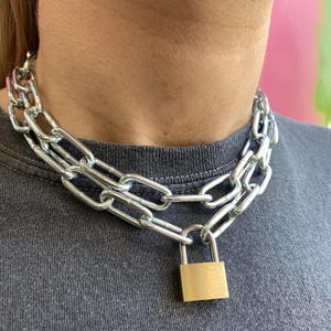 Può includere: Una collana a catena argentata con un ciondolo a forma di lucchetto dorato. La catena è a strati e ha un design a maglie grosse.