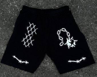 May include: Black shorts with white graphic designs. The shorts feature a barbed wire design at the bottom hem, a chain and spiked ball design, and a chain-link fence design. The shorts have a drawstring waistband.