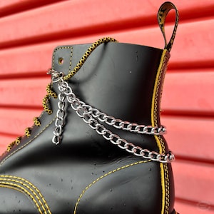 Op de afbeelding: Zwarte leren laars met gele stiksels en een zilveren kettingdetail. De laars heeft een vetersluiting aan de voorkant en een treklipje aan de achterkant.