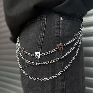 Puede incluir: Cinturón de cadena plateada con tres capas de cadenas. Cada cadena presenta pequeños dijes en forma de diente. El cinturón se usa sobre unos vaqueros negros.