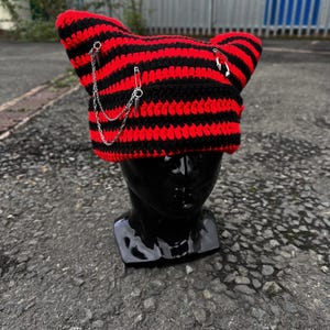 May include: A handmade crochet cat ear hat in red and black stripes. The hat is adorned with silver-colored safety pins and a chain. The hat is displayed on a black mannequin head.