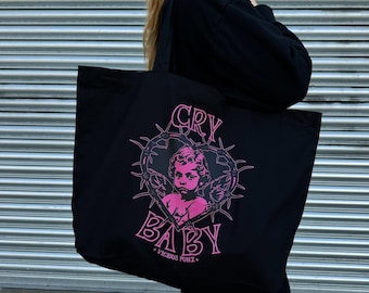 Puede incluir: Bolso tote negro con un gráfico rosa de un querubín dentro de un corazón de alambre de púas, con las palabras "CRY BABY" y "VICIOUS PUNX+". El bolso se muestra sobre un fondo de metal corrugado plateado.