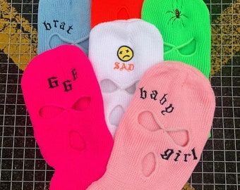 Puede incluir: Seis máscaras de esquí diferentes con varios diseños bordados. Las máscaras son de color rosa, azul, blanco, verde y naranja. Los diseños incluyen las palabras "brat", "66F", "sad", "baby girl" y una araña.