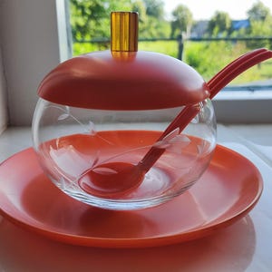 Könnte beinhalten: Eine Zuckerdose aus klarem Glas mit geätzten Blumenmustern, ausgestattet mit einem leuchtend orangefarbenen Kunststoffdeckel mit transparentem bernsteinfarbenem Griff, einer passenden orangefarbenen Untertasse und einem kleinen Löffel.