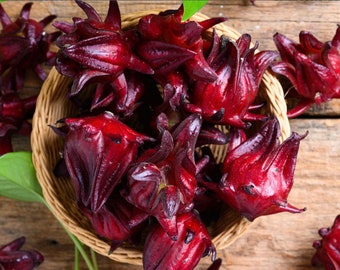 May include: A woven basket filled with vibrant red hibiscus flowers. The flowers have a deep burgundy hue and are arranged on a rustic wooden surface. Green leaves are visible, adding contrast to the scene.