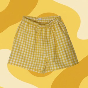 May include: Yellow and white checkered shorts with an elastic waistband. The shorts have two pockets and a small tag on the waistband. The background is a yellow and cream-colored abstract design.