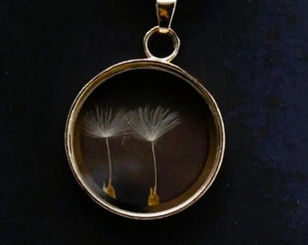 May include: A gold pendant necklace with a round glass locket featuring two delicate dandelion seeds inside. The locket is suspended from a black cord.