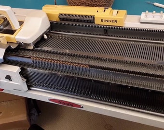 May include: A vintage Singer knitting machine in cream and yellow. The machine has a series of metal needles and a gauge dial. The word "SINGER" is printed on a yellow component. The machine is on a white surface.