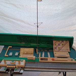 Peut inclure: Machine à tricoter Brother-Knit vintage, vert d'eau et beige, avec un coffret de rangement vert. La machine comporte des boutons et des leviers, et un livret de motifs est visible. Un grand support métallique est fixé.