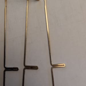 以下が含まれることがあります： ラグフックやラッチフックプロジェクトに使用される、3つの金属製ラッチフックと金色の針のクローズアップ。フックは銀色で、上部に湾曲したフックと曲がったハンドルが付いています。針は金色です。
