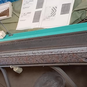 May include: A vintage knitting machine with a teal and cream color scheme. The machine has a long bed with numerous needles, a cream-colored control panel, and various accessories, including a needle selector and instruction manual.