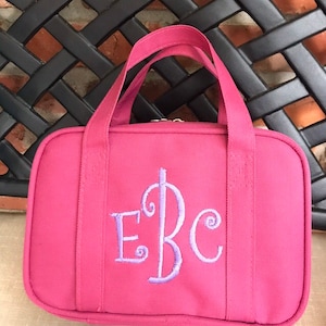May include: A pink travel bag with a top handle and a zipper closure. The bag features an embroidered monogram in light purple. The bag is made of a durable fabric and is designed for carrying toiletries or other small items.