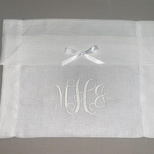 May include: A white rectangular fabric case featuring a folded top flap secured with a small white satin ribbon bow. The center of the case is decorated with an embroidered white monogram featuring the stylized cursive letters M, H, and J.