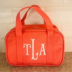 May include: Coral-colored duffel bag with white embroidered initials "TLA". The bag has two short handles and a zipper closure. The bag is made of a durable fabric and is suitable for travel or everyday use.