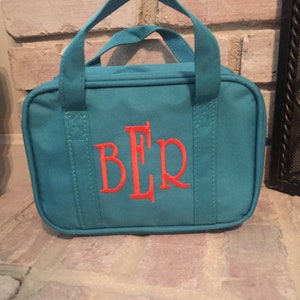 May include: Teal canvas tote bag with a top handle and the embroidered initials "BLR" in coral. The bag has a rectangular shape and is sitting on a brick surface.