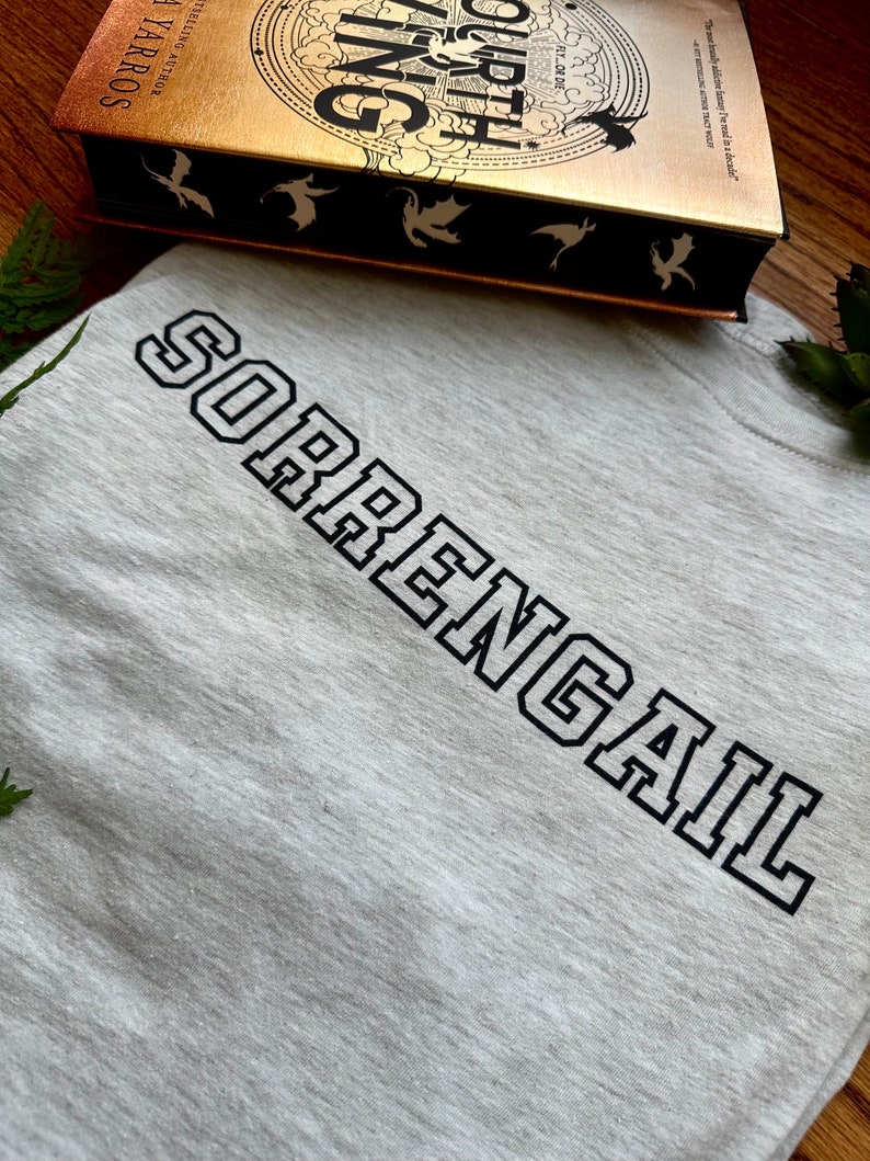 K&ouml;nnte beinhalten: Ein graues T-Shirt mit dem Text "SORRENGAIL" in schwarzen umrandeten Buchstaben. Das T-Shirt liegt auf einer Oberfl&auml;che, im Hintergrund ist ein Buch mit dem Titel "Fourth Wing" zu sehen.