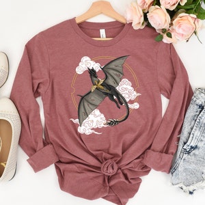 Puede incluir: Una camiseta de manga larga de color granate con un gráfico de un dragón negro volando en un círculo con nubes y detalles dorados.