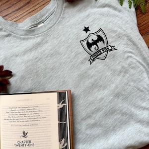 K&ouml;nnte beinhalten: Ein graues T-Shirt mit einer schwarzen Grafik eines Drachen und eines Sterns auf einem Schild mit dem Text "Fourth Wing". Das T-Shirt liegt auf einer Holzoberfl&auml;che neben einem ge&ouml;ffneten Buch mit dem Text "Chapter Twenty-One" auf dem Cover.