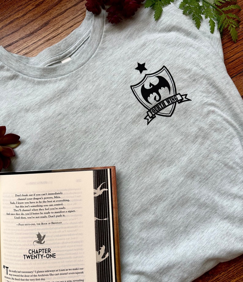 K&ouml;nnte beinhalten: Ein graues T-Shirt mit einer schwarzen Grafik eines Drachen und eines Sterns auf einem Schild mit dem Text "Fourth Wing". Das T-Shirt liegt auf einer Holzoberfl&auml;che neben einem ge&ouml;ffneten Buch mit dem Text "Chapter Twenty-One" auf dem Cover.