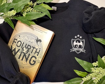 May include: A black sweatshirt with a white embroidered crest featuring a dragon and the text "Fourth Wing". A book titled "Fourth Wing" with a dragon design is partially visible in the bottom left corner.