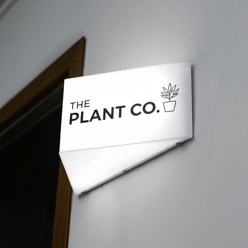 May include: A white, rectangular sign with the text "THE PLANT CO." in black, along with a small potted plant graphic. The sign is mounted on a gray wall, with a door frame visible in the background.