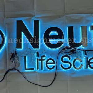 May include: A custom-made illuminated sign with the text "Neutra Life Sciences" in black letters with a blue halo. The sign is made of stainless steel and has a modern design.