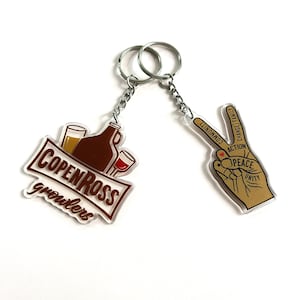 May include: Two keychains, one with a brown and white logo that says "COPENROSS growlers" and a picture of a beer bottle and glasses, and the other with a peace sign with the words "PEACE UNITY ACTION CHANGE FAMILY COMMUNITY" around it.
