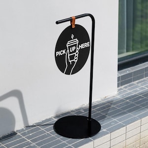 May include: A black metal sign with a circular design that reads "PICK UP HERE" in white. The sign features a hand holding a coffee cup graphic. The sign is attached to a black metal stand with a round base and a curved top.