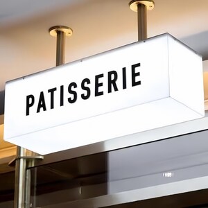 May include: A rectangular, illuminated sign with the word "PATISSERIE" in bold, black capital letters. The sign is white and hangs from the ceiling by two brass-colored rods.