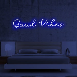 May include: A blue neon sign with white lettering that reads "Good Vibes" hanging on a dark gray concrete wall above a bed with white bedding.