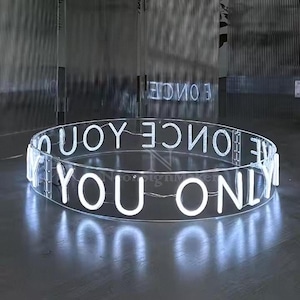 Puede incluir: Un letrero de neón circular con las palabras "YOU ONLY ONCE" en letras blancas iluminadas. El letrero está hecho de acrílico transparente y está iluminado con luces LED blancas brillantes. El letrero está sobre una superficie oscura.