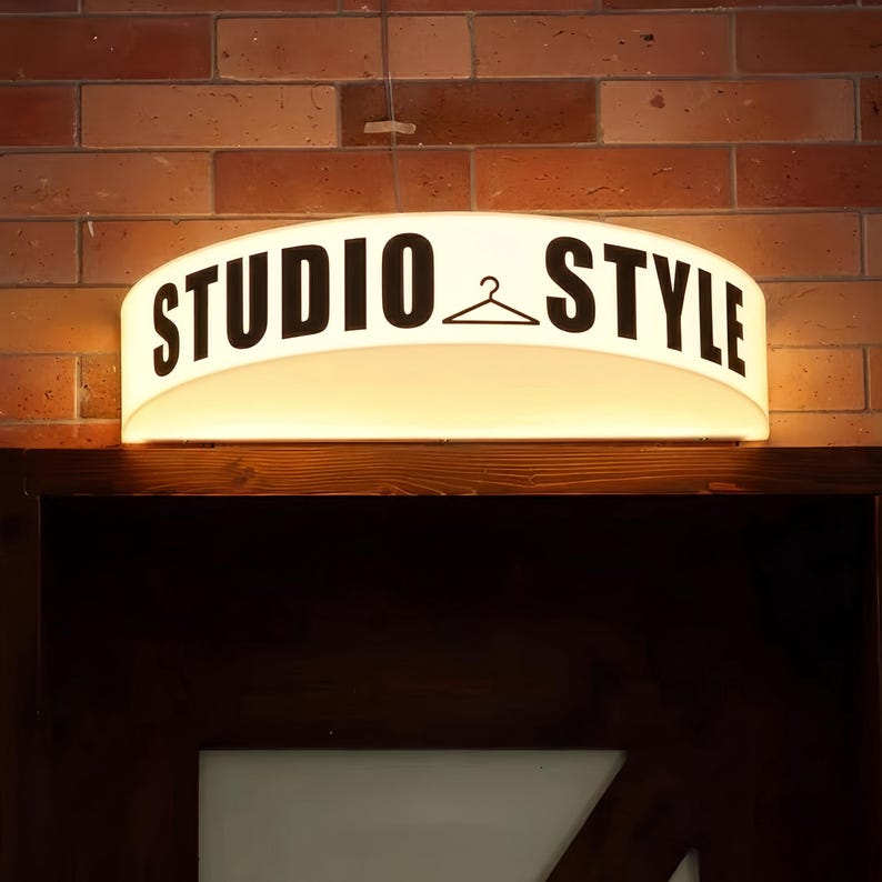 May include: A backlit sign with the words "STUDIO & STYLE" in black, with a hanger symbol between the words. The sign is mounted above a dark wooden door frame against a brick wall.
