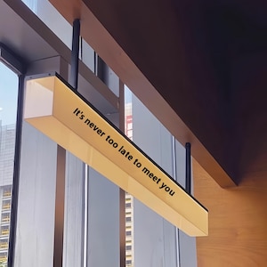 May include: A rectangular, illuminated sign with the text "It's never too late to meet you" in black. The sign is suspended from the ceiling with black rods. The sign has a yellow glow, and is near a window.
